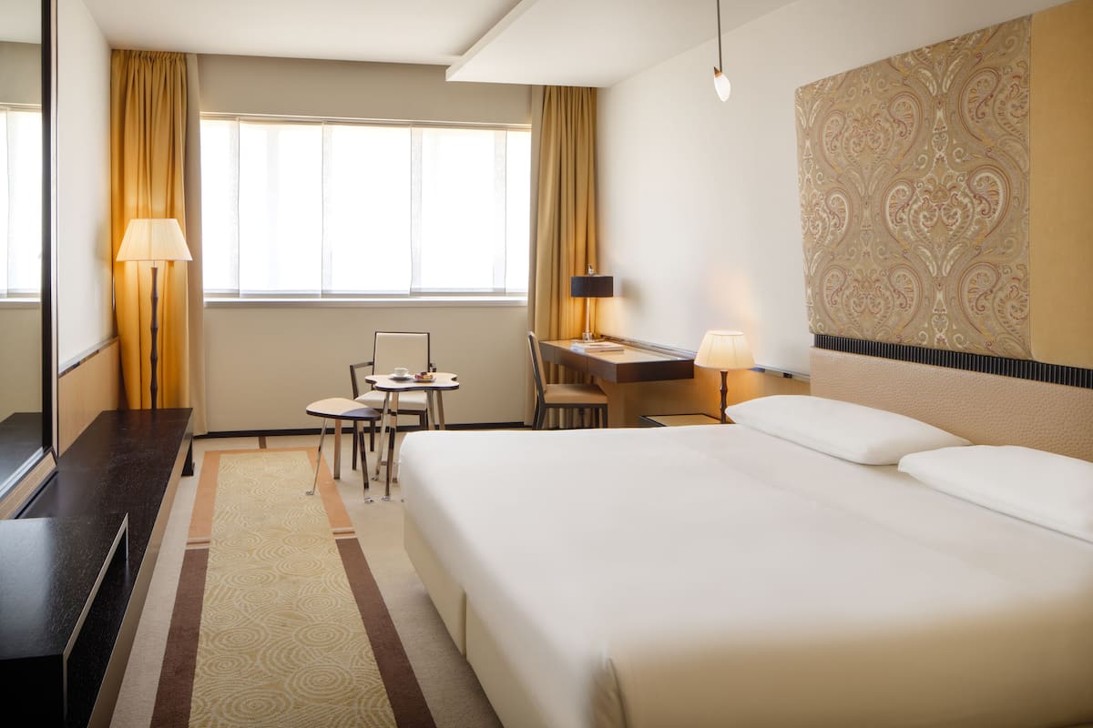 Una habitación de hotel con una cama grande, un escritorio con una silla, una mesita pequeña y una lámpara.