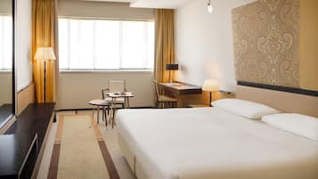 A hotel room with a large bed, a desk with a chair, a small table, and a lamp.