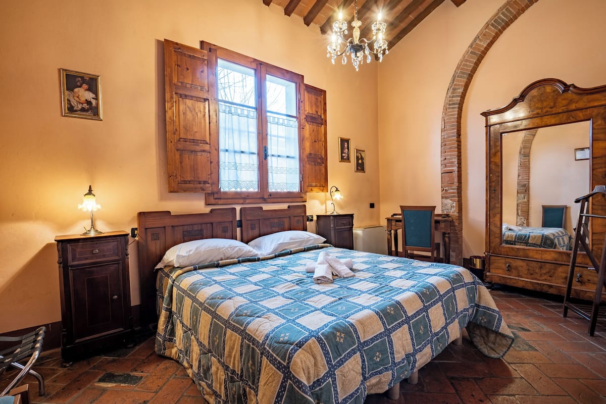 A bedroom with a large bed, wooden headboard, a window with shutters, a chandelier, a mirror, and a dresser.