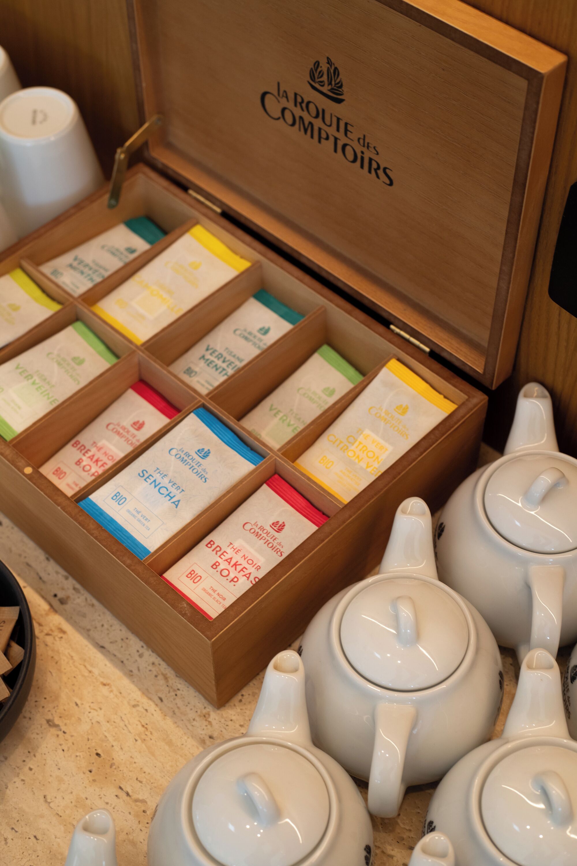 Une boîte en bois étiquetée « La Route des Comptoirs » contenant des sachets de thé et des théières blanches.
