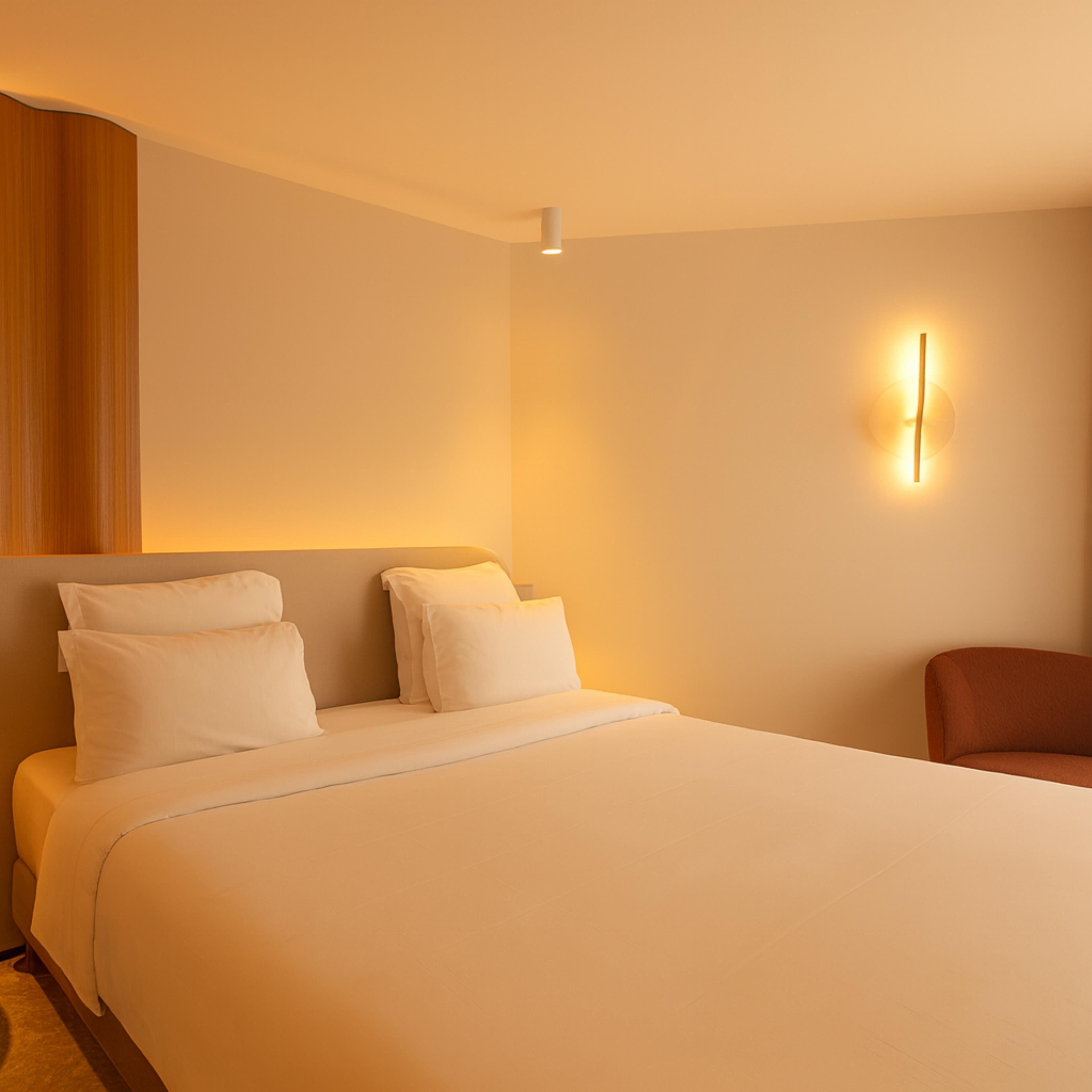 A hotel room with a large bed, a wooden headboard, a wall-mounted light, and a brown armchair.