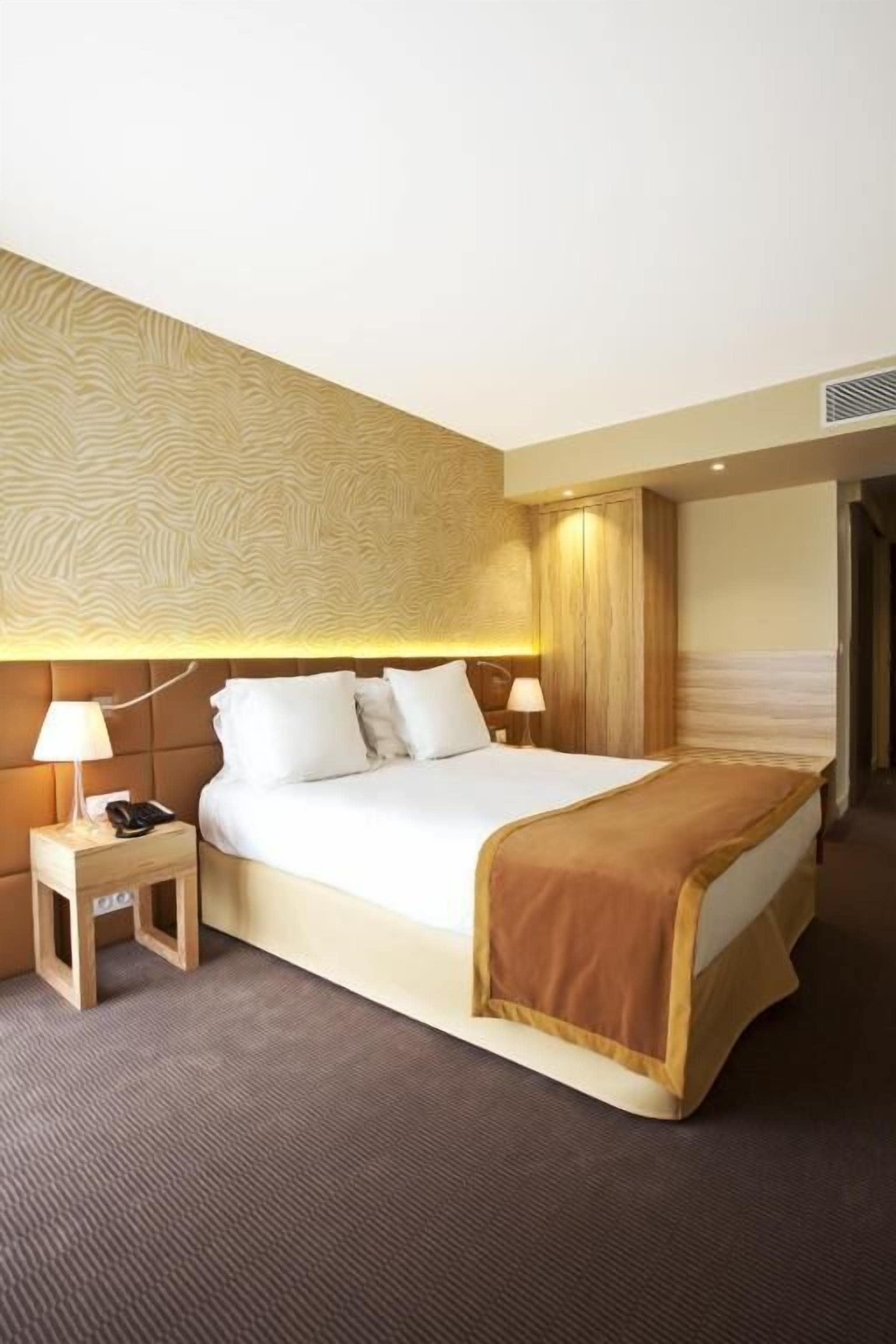 A hotel room with a large bed, two bedside tables, a headboard with a textured design, and a carpeted floor.