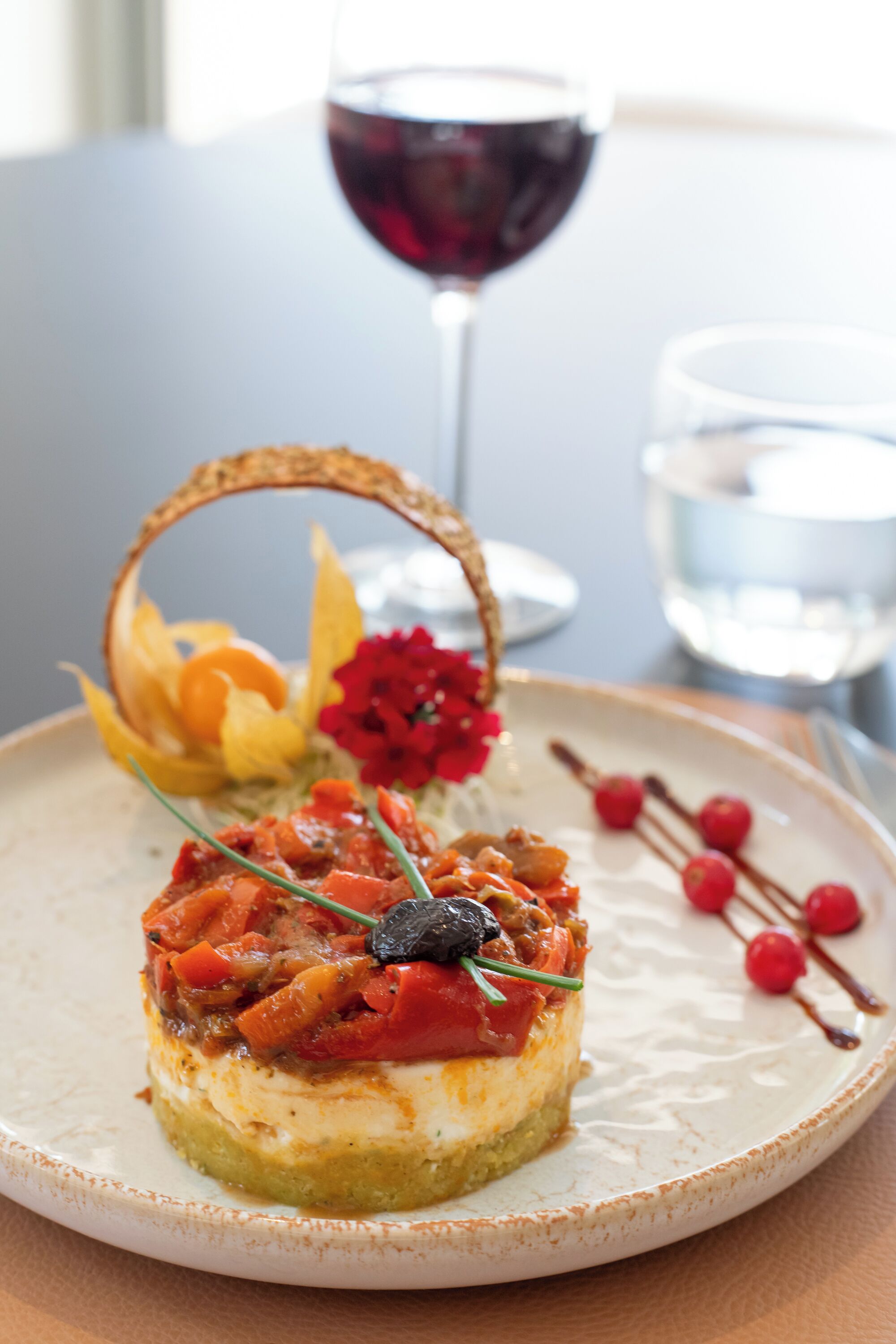 A dessert plate with a small cake topped with red sauce and a black olive, accompanied by a glass of red wine and a glass of water.