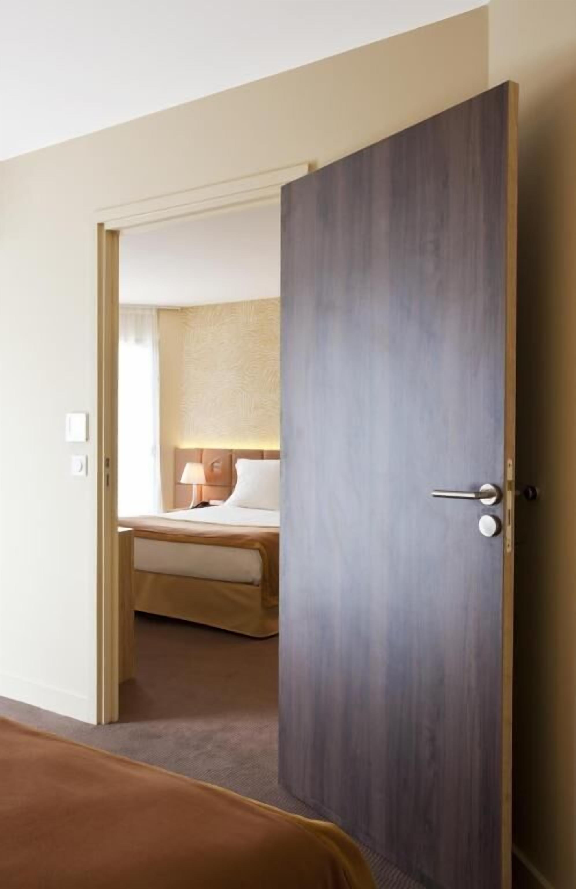 A hotel room with a wooden door, a bed with a white headboard, and a bedside lamp.