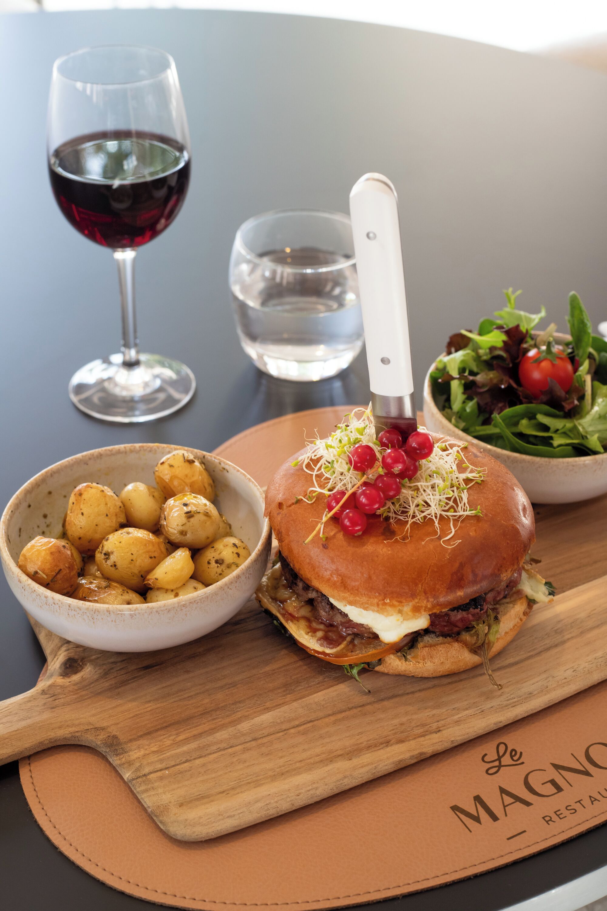 Un repas composé d’un hamburger, de pommes de terre rôties et d’une salade, le tout présenté sur une planche en bois, avec un verre à vin et un verre à eau en arrière-plan.