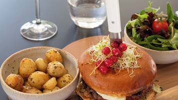 Un repas composé d’un hamburger, de pommes de terre rôties et d’une salade, le tout présenté sur une planche en bois, avec un verre à vin et un verre à eau en arrière-plan.