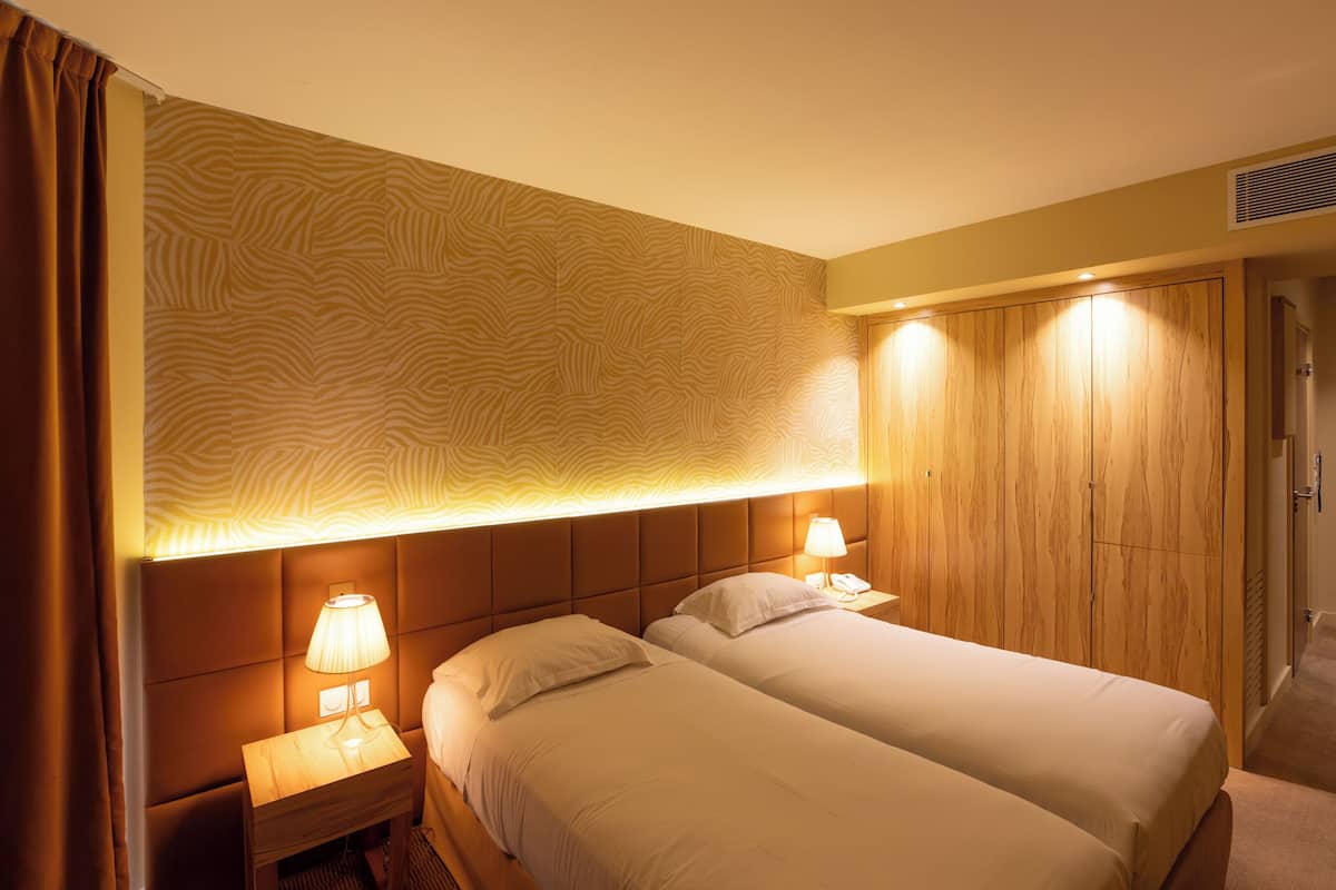 A hotel room with two beds, a headboard with a patterned design, bedside lamps, and a wooden wardrobe.