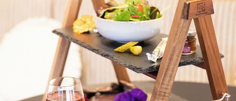 Un soporte de madera con un tazón de ensalada, un vaso de bebida roja y algunos alimentos sobre una superficie oscura.