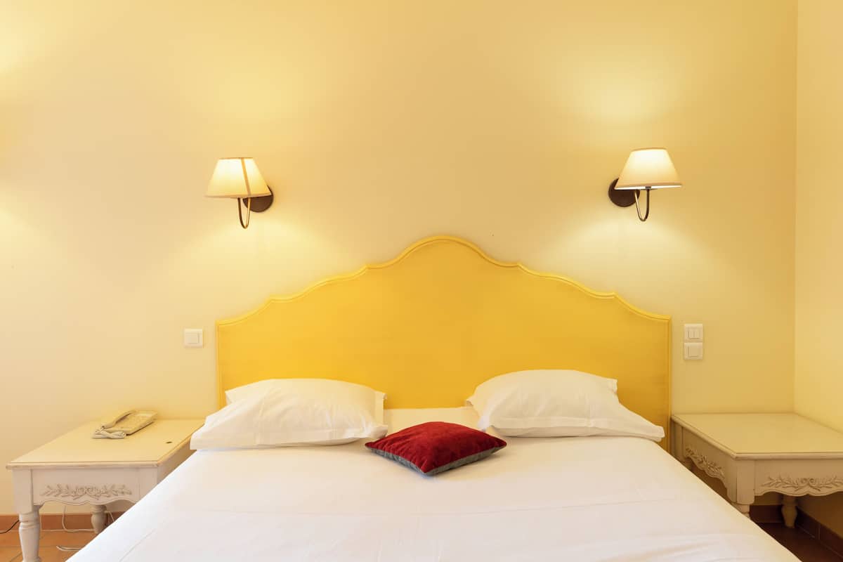 A hotel room with a bed, two bedside lamps, a nightstand with a phone, and a red pillow.