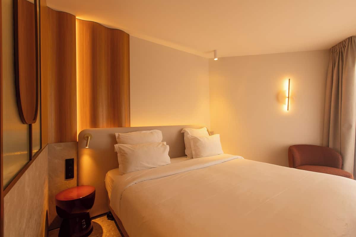 A hotel room with a bed, a chair, a wall-mounted light, and a toilet.