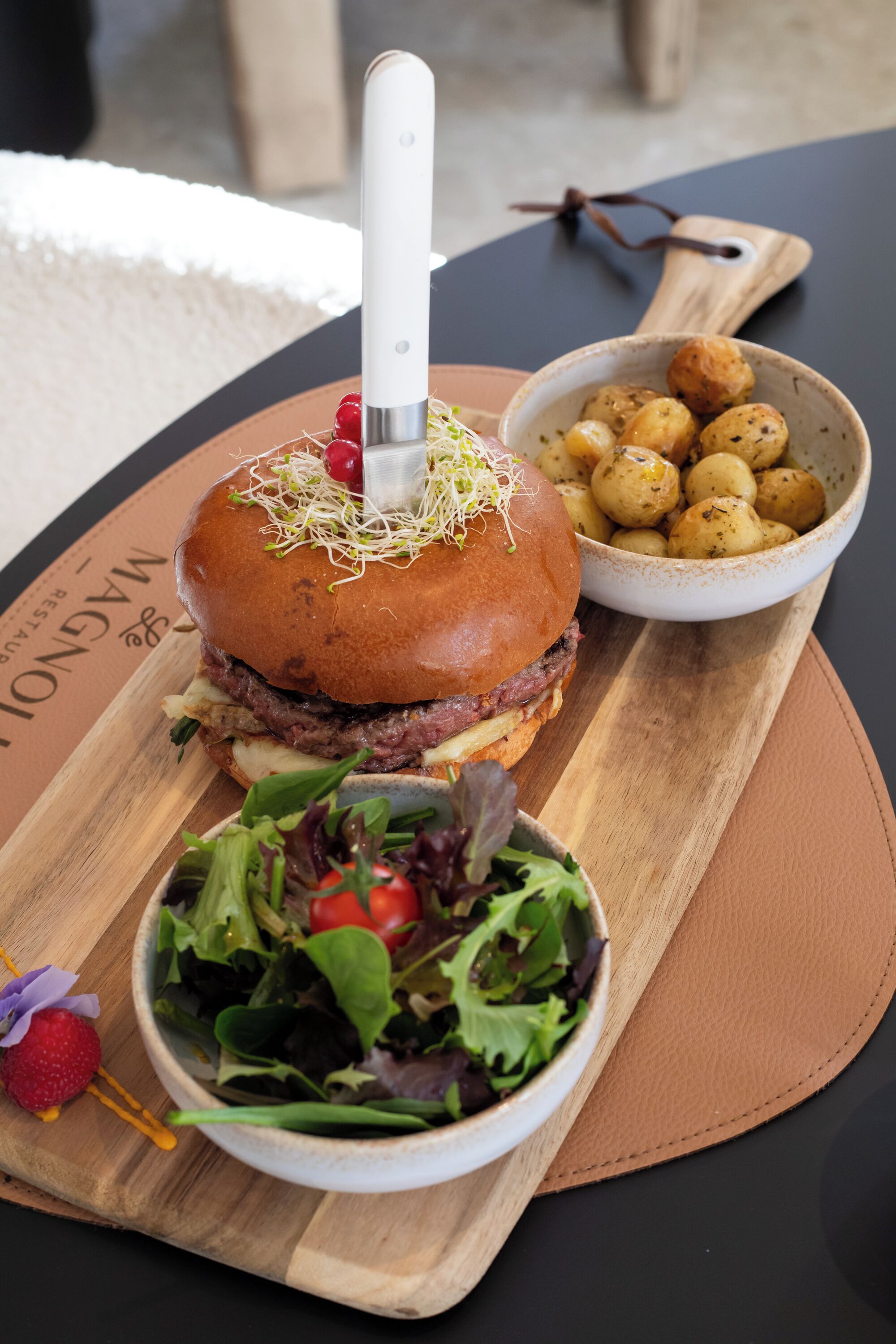 A burger with a knife, a bowl of potatoes, and a salad on a wooden board.