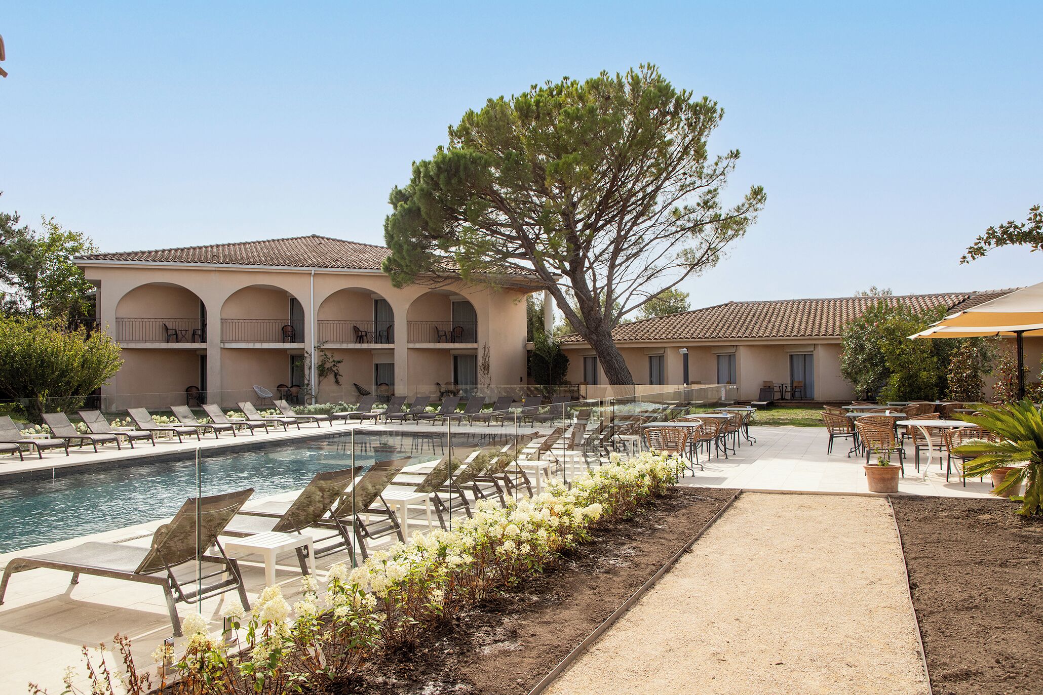 A pool area with lounge chairs, a building with arches, and a large tree.