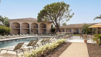 Un espace piscine avec des chaises longues, un bâtiment orné d’arches et un grand arbre.