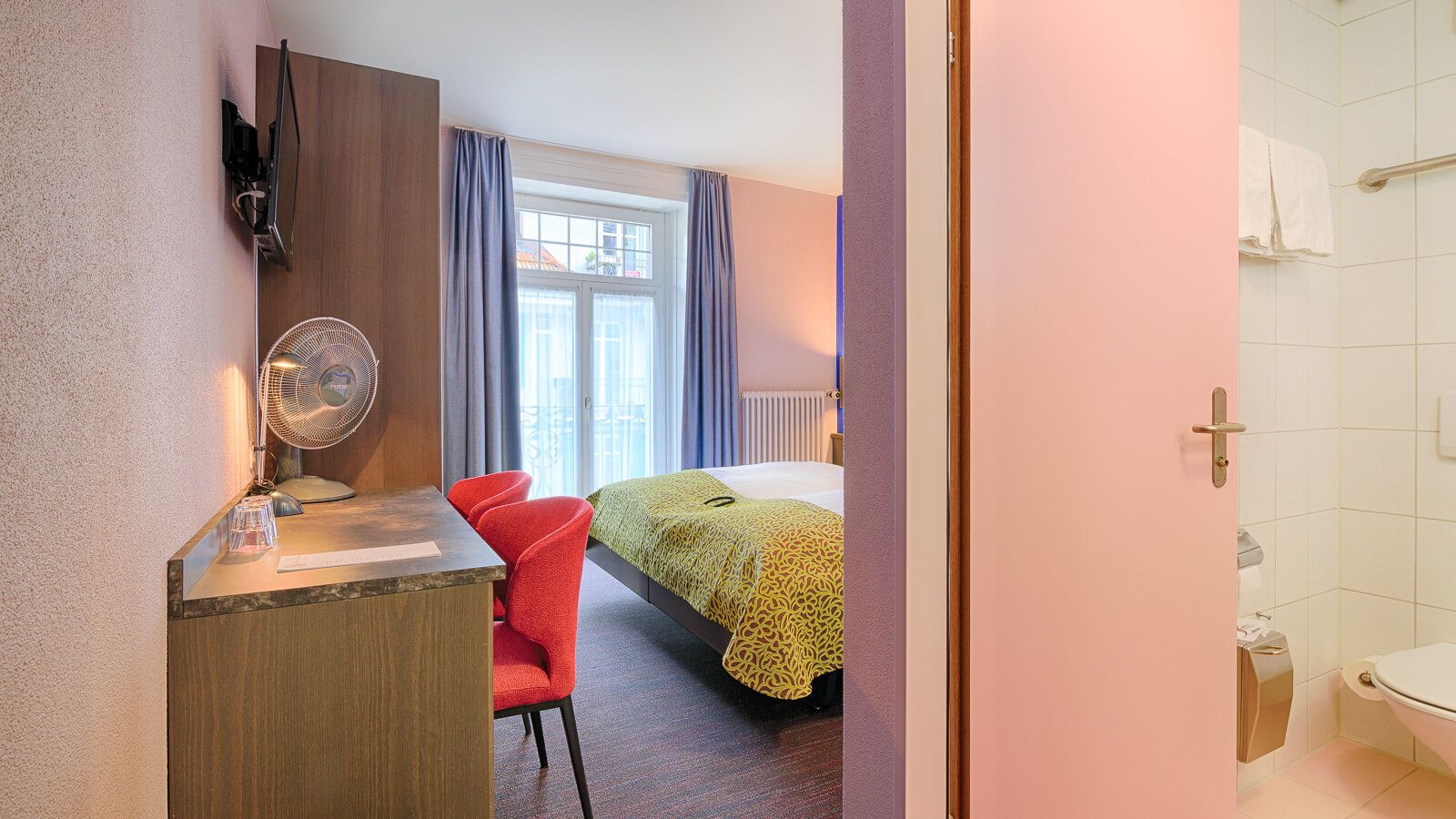 Habitació d'hotel amb un llit, una taula de treball amb una cadira, un ventilador i vistes a l'exterior a través d'una finestra amb cortines.