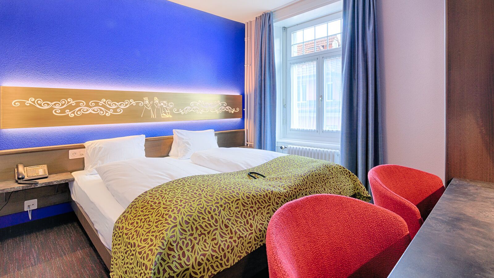 Una habitación de hotel con cama, mesita de noche con teléfono, dos sillas rojas, ventana con cortinas y pared azul con un panel decorativo.