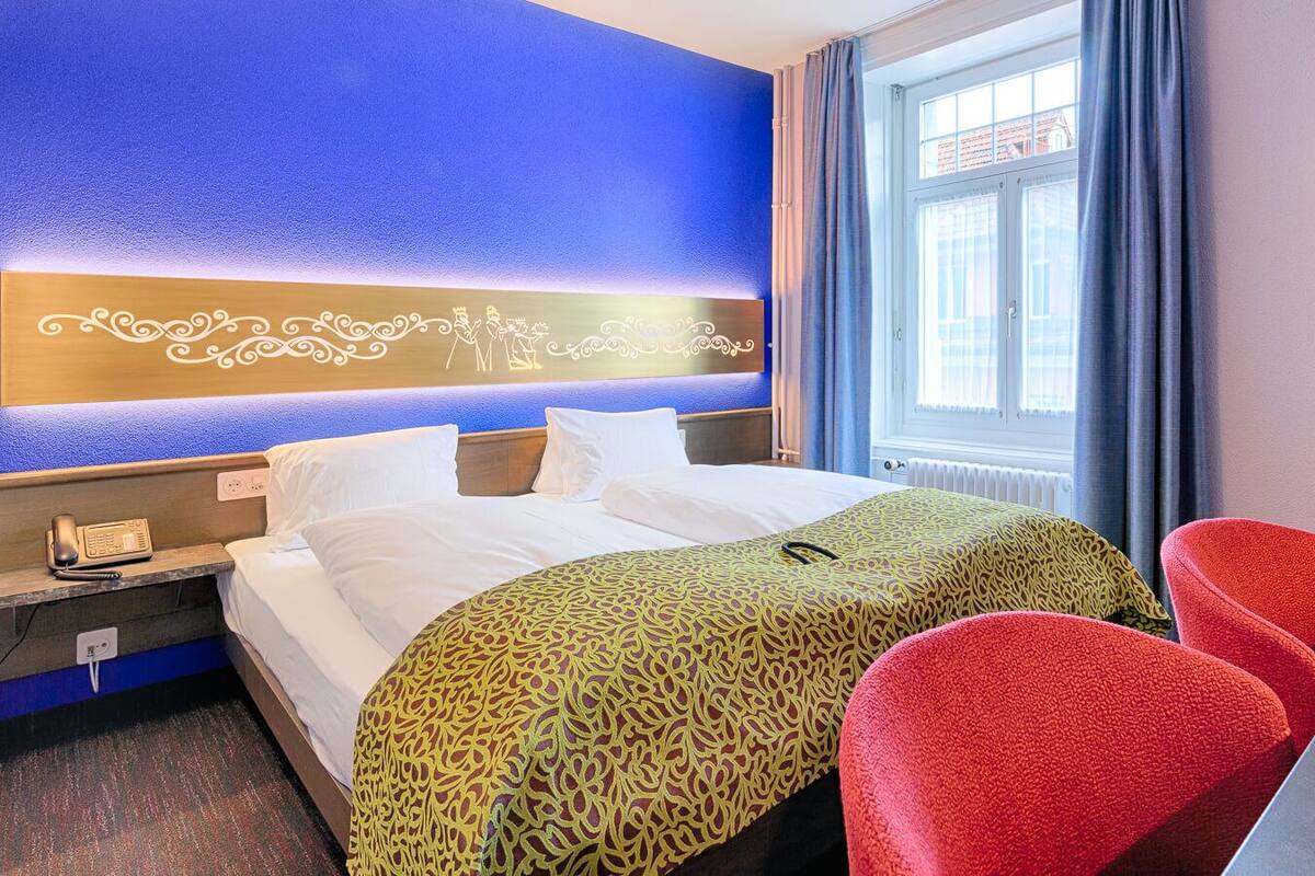 Una habitación de hotel con cama, mesita de noche con teléfono, dos sillas rojas, ventana con cortinas y pared azul con un panel decorativo.