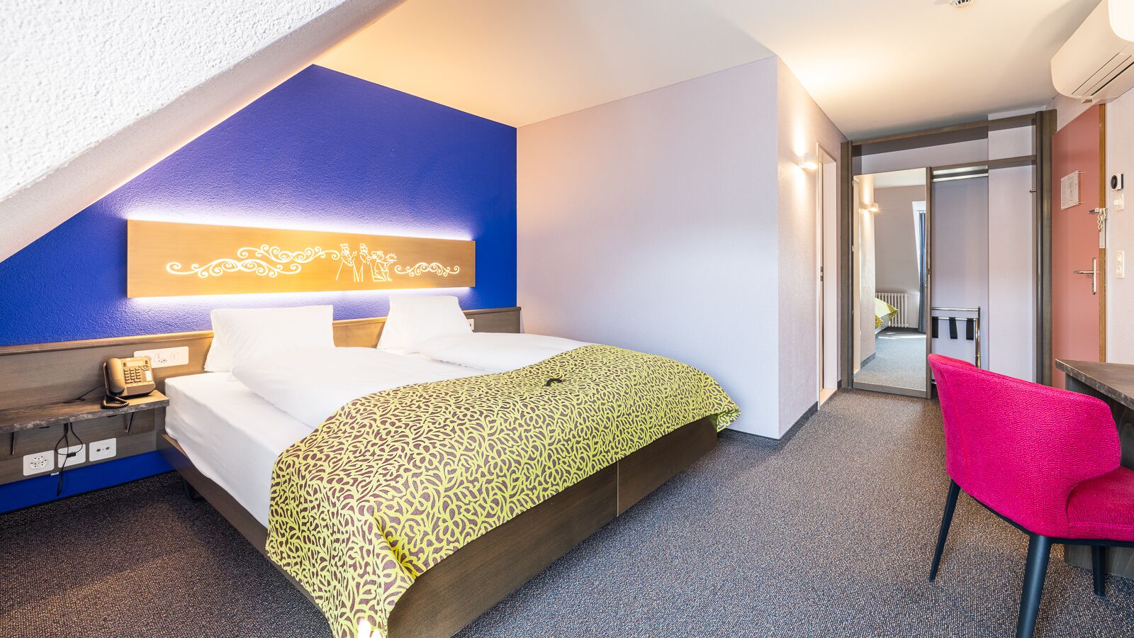 A hotel room with a bed, a desk, a chair, and a phone on the nightstand.