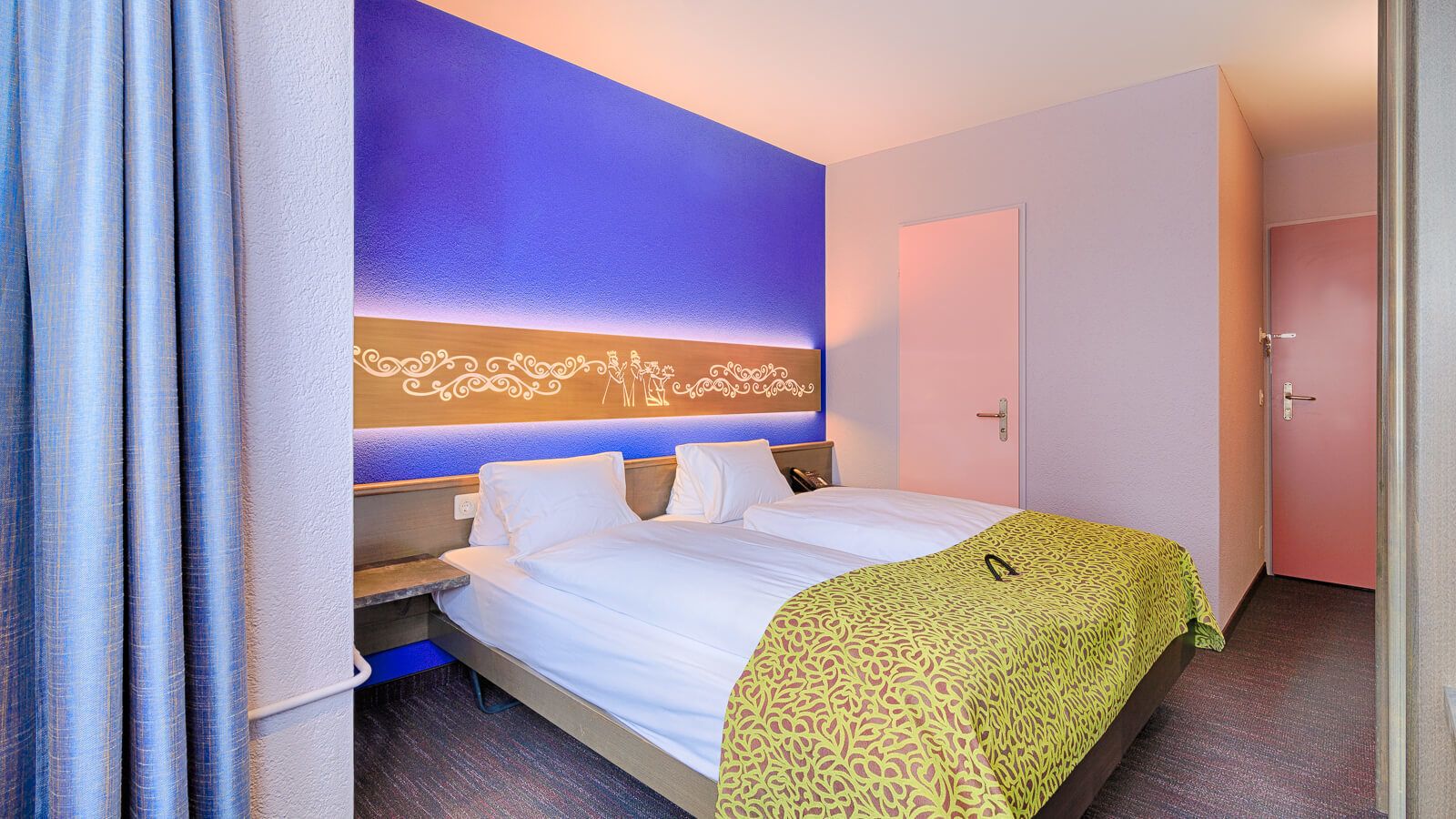 A hotel room with a bed, a nightstand, a wall-mounted light, and a door.