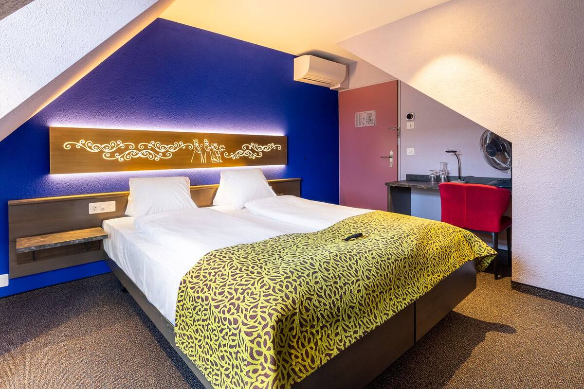Ett hotellrum med en säng, ett sängbord, en röd stol och ett litet pentry.