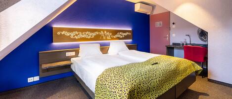 A hotel room with a bed, a bedside table, a red chair, and a small kitchenette.