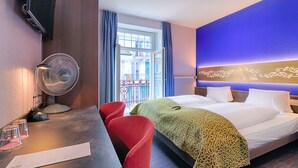 Quarto de hotel com cama, escrivaninha com ventoinha, cadeiras vermelhas e vista para o exterior.