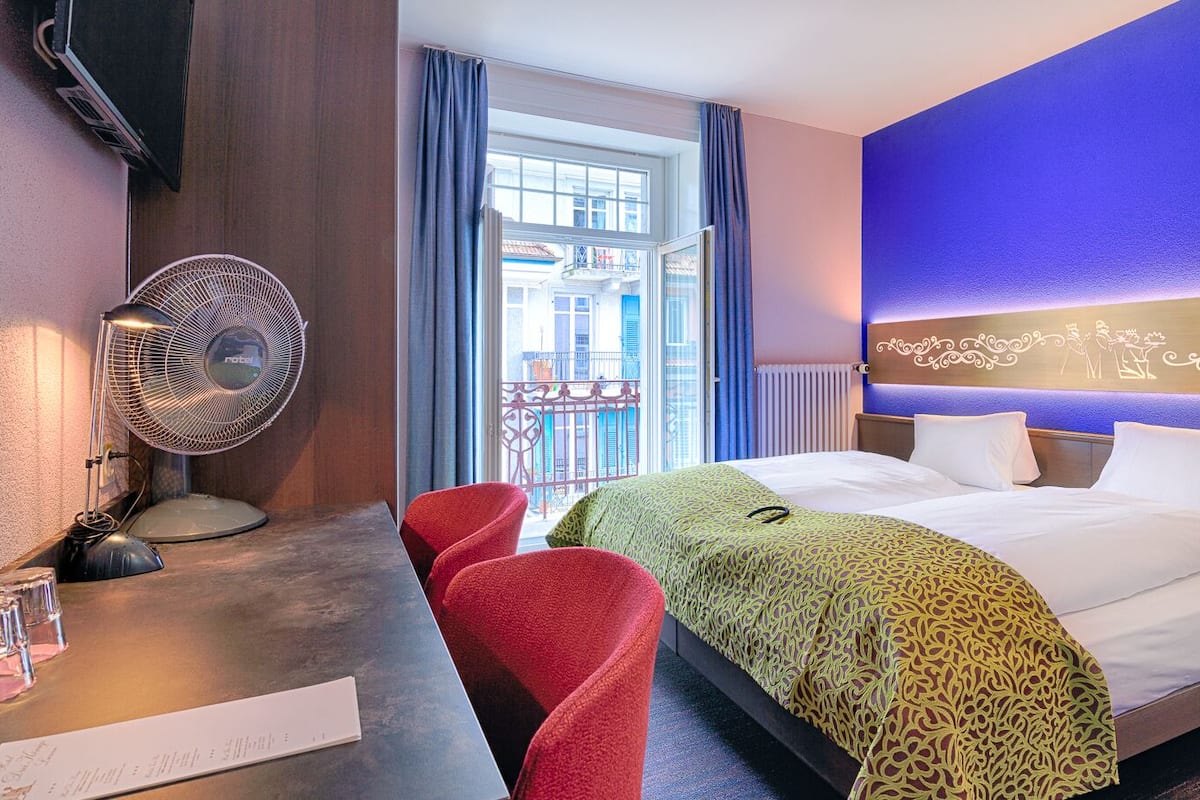 Ett hotellrum med en säng, ett skrivbord med en fläkt, röda stolar och utsikt över omgivningen.