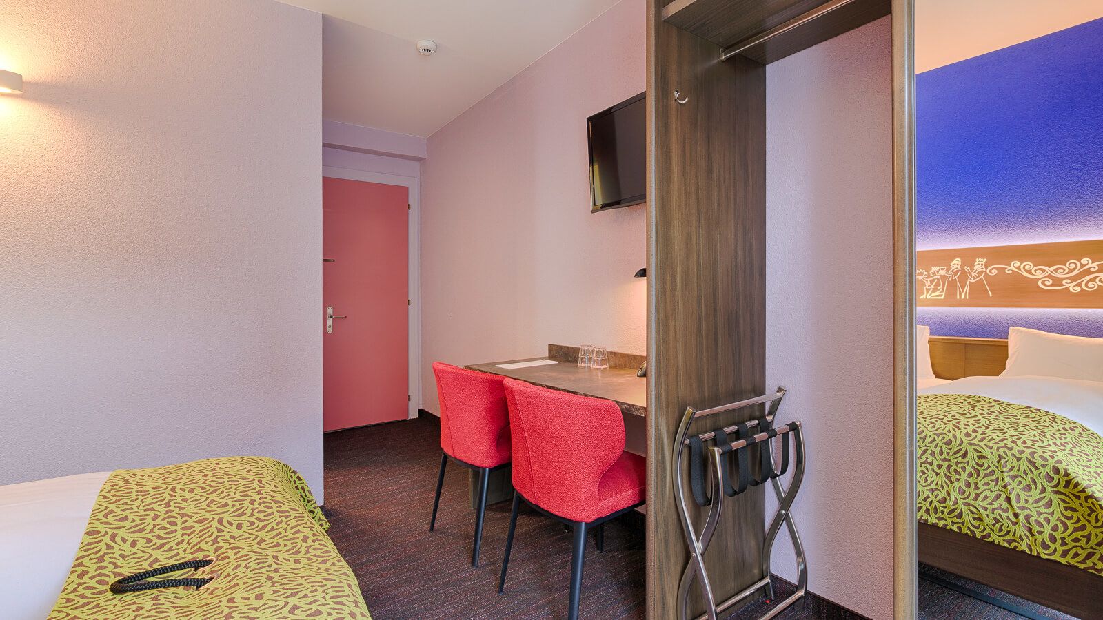 Une chambre d’hôtel avec deux lits, un bureau avec deux chaises rouges, un téléviseur fixé au mur et une porte donnant sur une autre pièce.