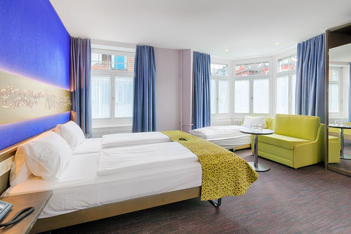 Ett hotellrum med två sängar, en gul fåtölj, ett litet bord och ett stort fönster med gardiner.