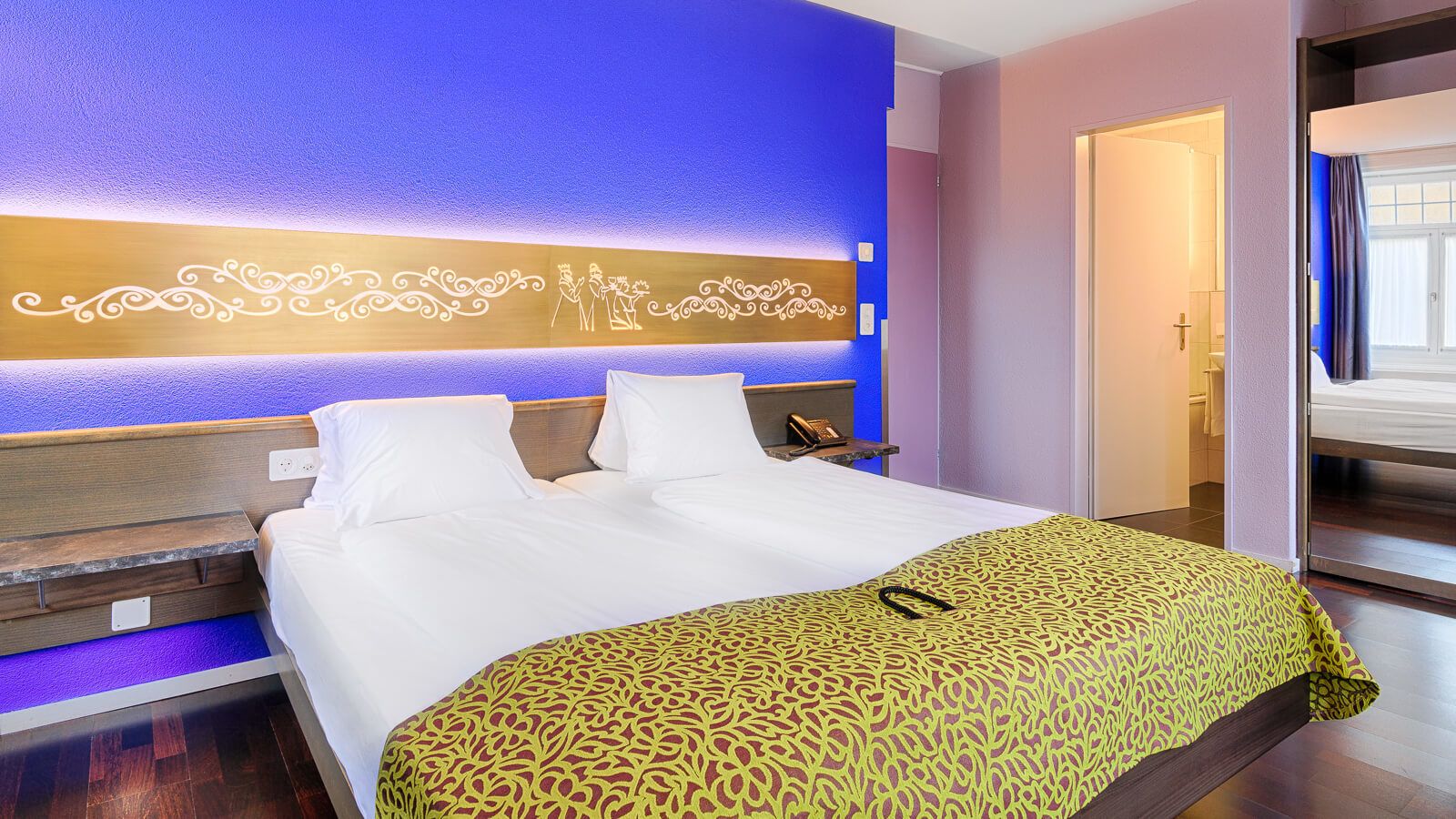 A hotel room with a large bed, a bedside table, a nightstand, and a wall with decorative lighting.