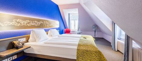 Quarto de hotel moderno com cama, mesa de cabeceira com telefone, pequena mesa com ventoinha e janela com cortinas.
