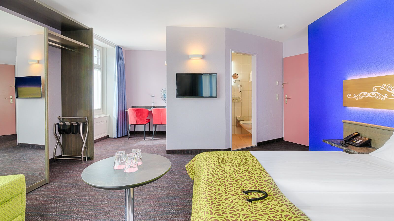 A hotel room with a bed, a small table with glasses, a TV, a bathroom, and a seating area with chairs.