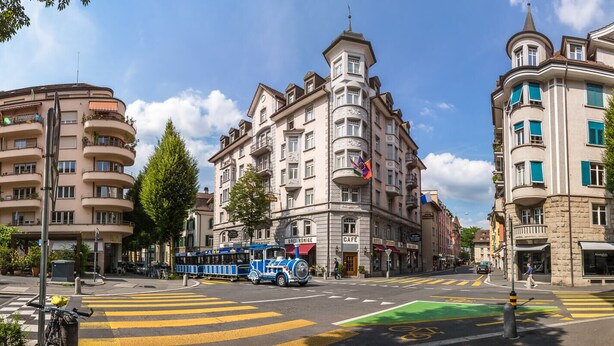 Uma rua com bondinho azul, edifícios históricos e faixa de pedestres.
