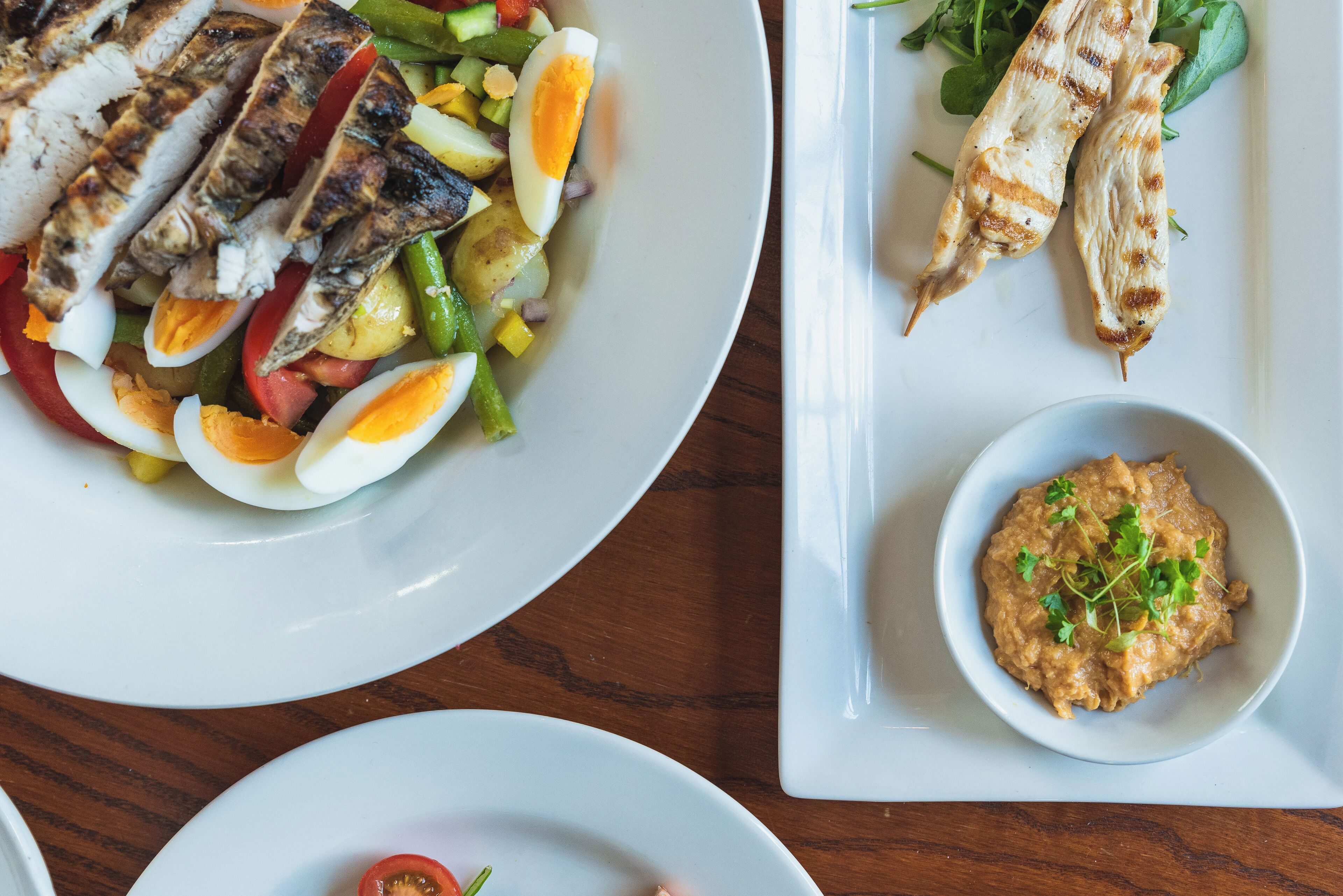 A table with three plates of food, including grilled chicken, vegetables, and a bowl of dip.