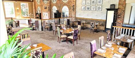 A spacious restaurant with high ceilings, exposed beams, and brick walls. Tables are set with white tablecloths and wine glasses.