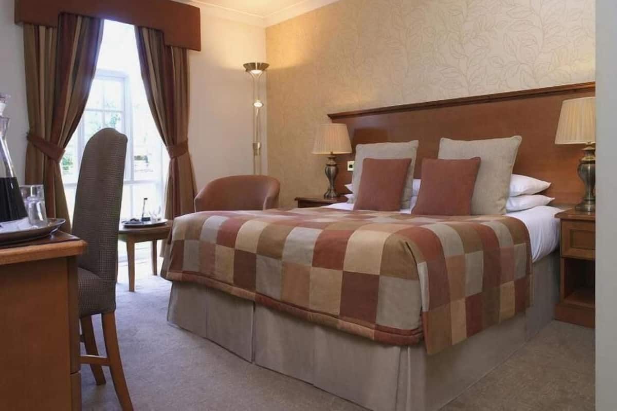 A hotel room with a bed, two bedside tables, a desk with a chair, a lamp, and a window with curtains.