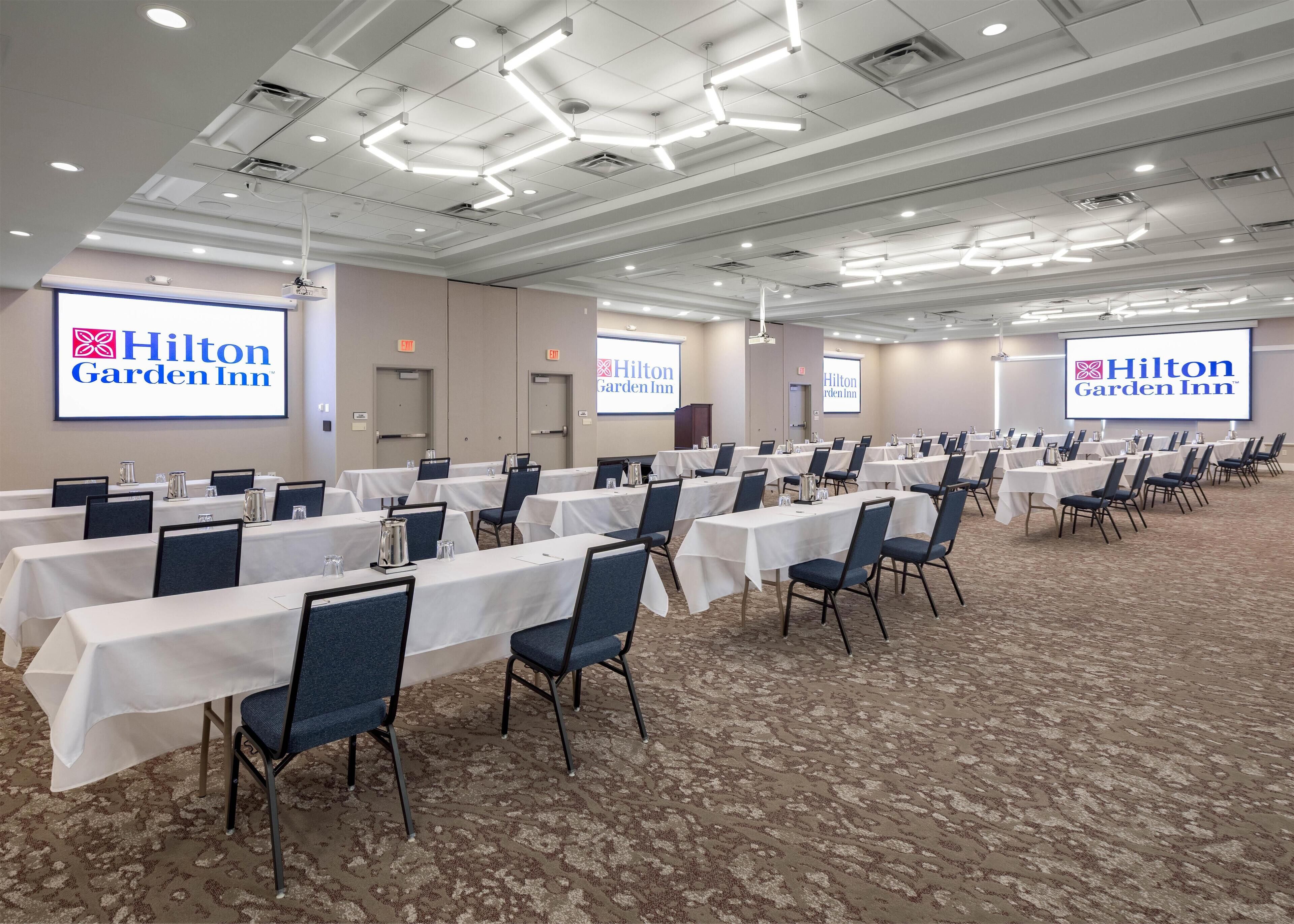 Una sala de conferencias en Hilton Garden Inn con varias mesas, sillas y pantallas de presentación.