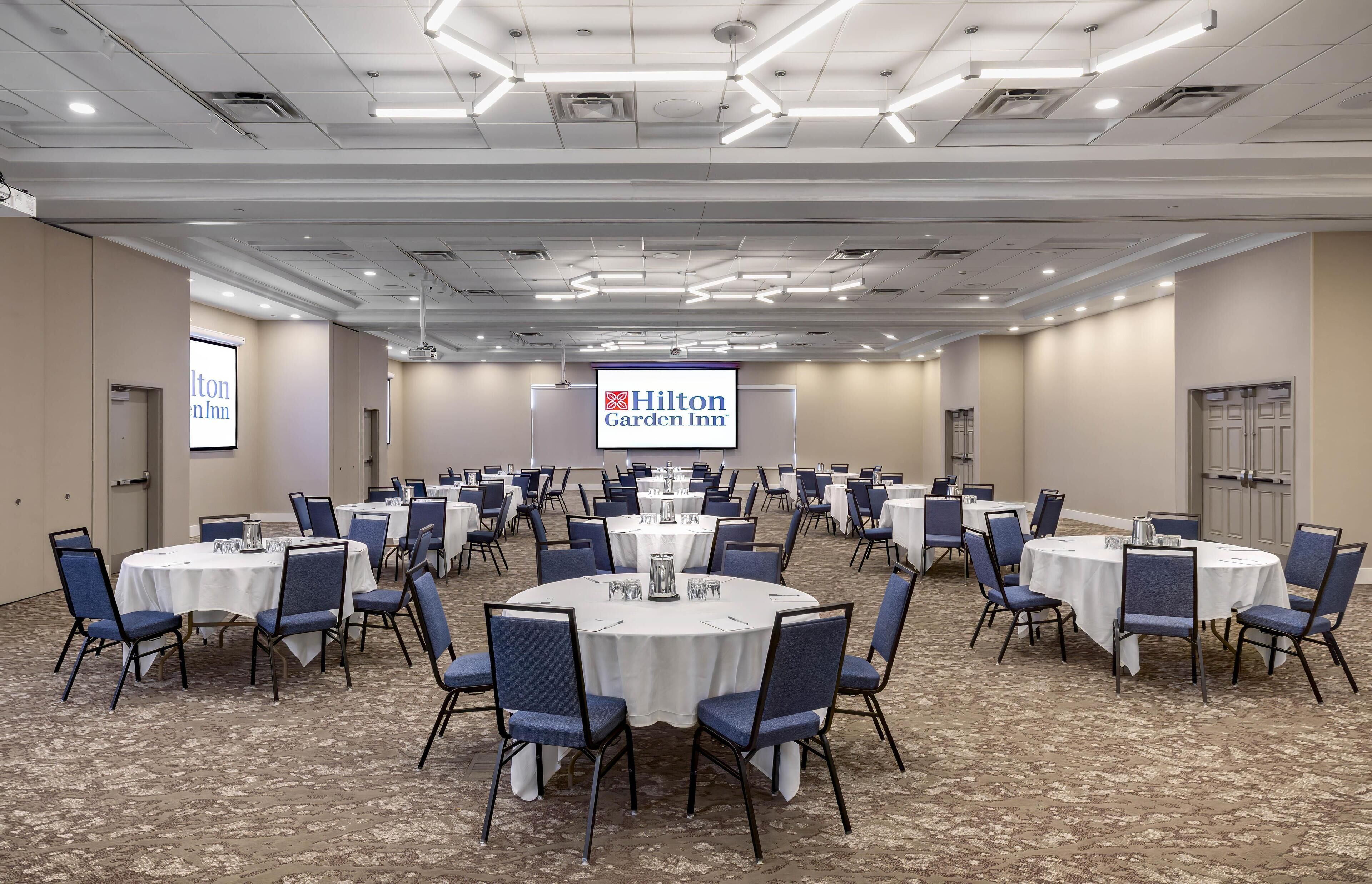 A conference room with multiple round tables, blue chairs, and a Hilton Garden Inn sign.