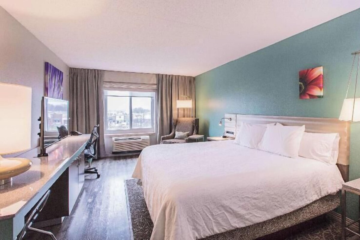 A hotel room with a large bed, a desk, a chair, a TV, and a window with curtains.