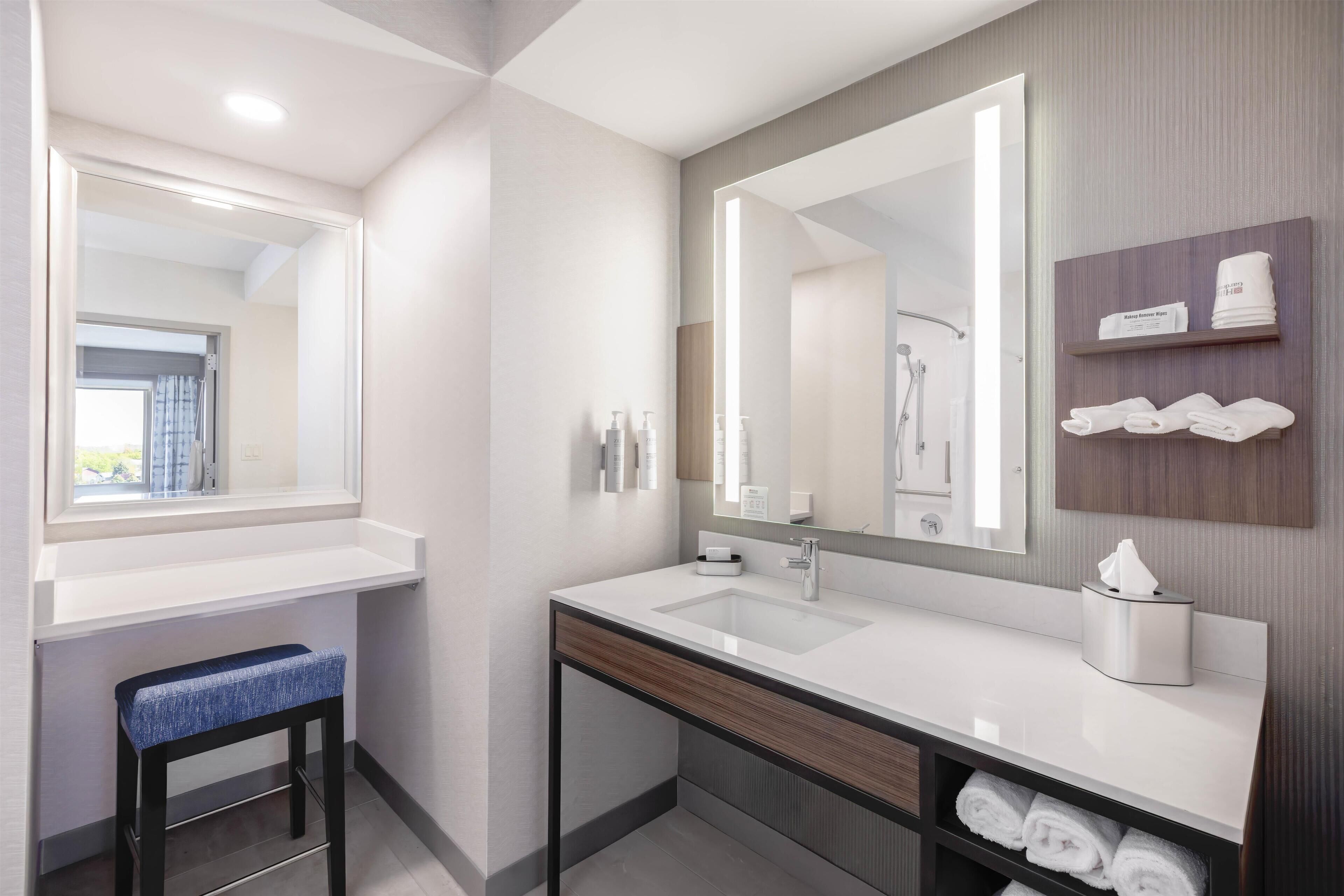A modern bathroom with a large mirror, two sinks, a towel rack, and a bench.