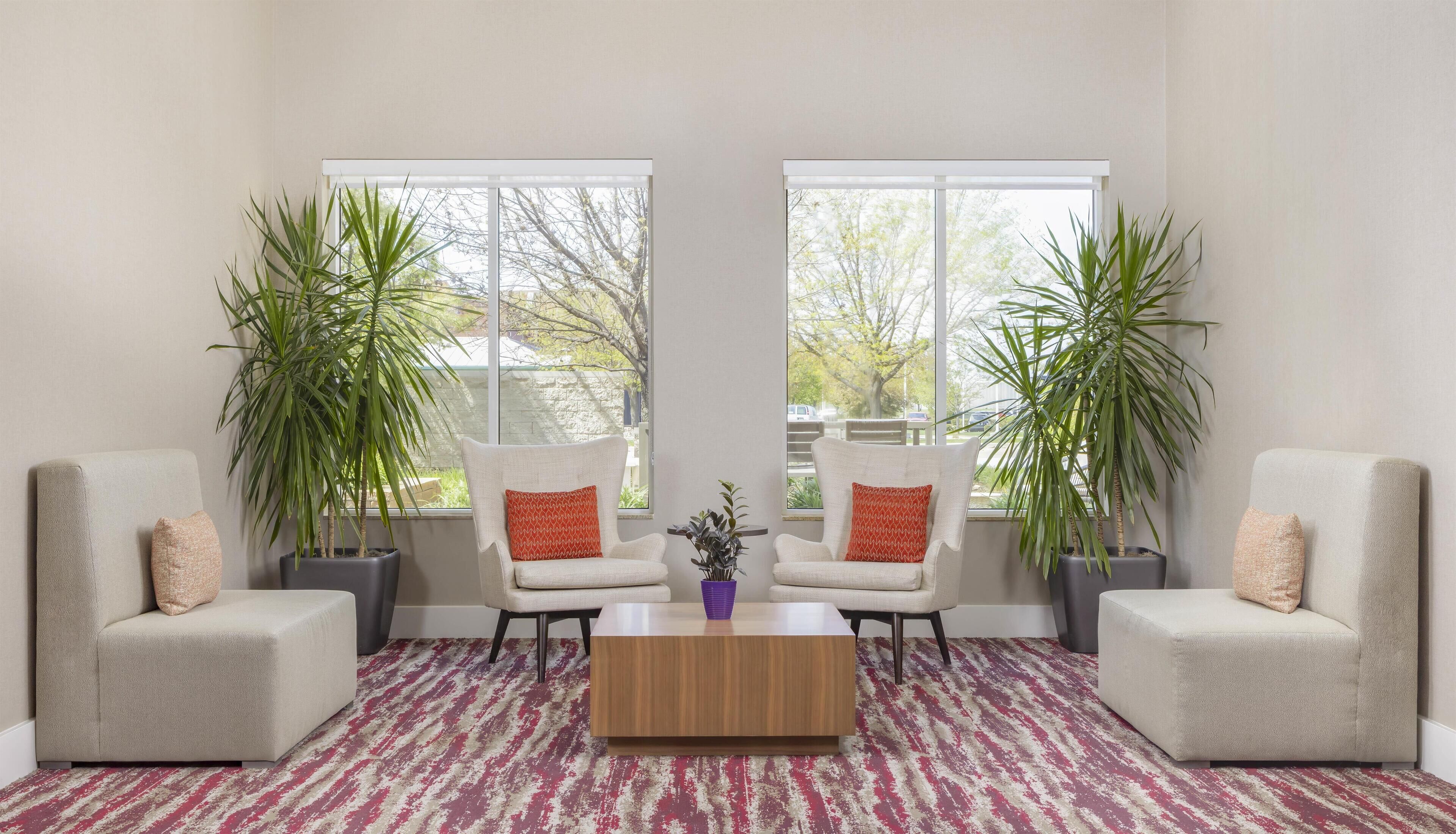 Un salón con dos sillones blancos, una mesa de centro de madera y ventanas amplias con plantas en macetas.