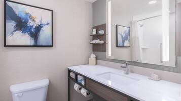 A modern bathroom with a white toilet, a sink, and a large mirror. There are shelves with toiletries and a framed abstract painting on the wall.