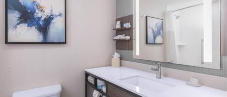 A modern bathroom with a white toilet, a sink, and a large mirror. There are shelves with toiletries and a framed abstract painting on the wall.