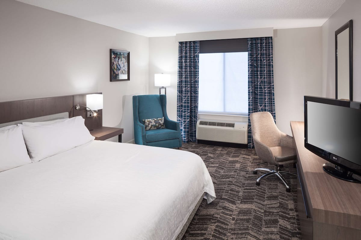 Een hotelkamer met een groot bed, een bureau met een flatscreen televisie, een blauwgroene fauteuil, een beige fauteuil en een raam met behang gordijnen.