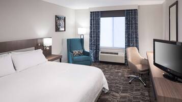 A hotel room with a large bed, a desk with a flat-screen TV, a teal armchair, a beige armchair, and a window with patterned curtains.