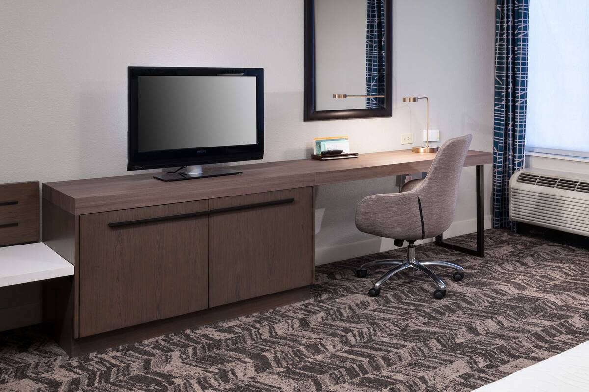 Hotelkamer met een houten bureau, een flatscreen televisie, een moderne stoel, een grote spiegel en een raam met gordijnen.