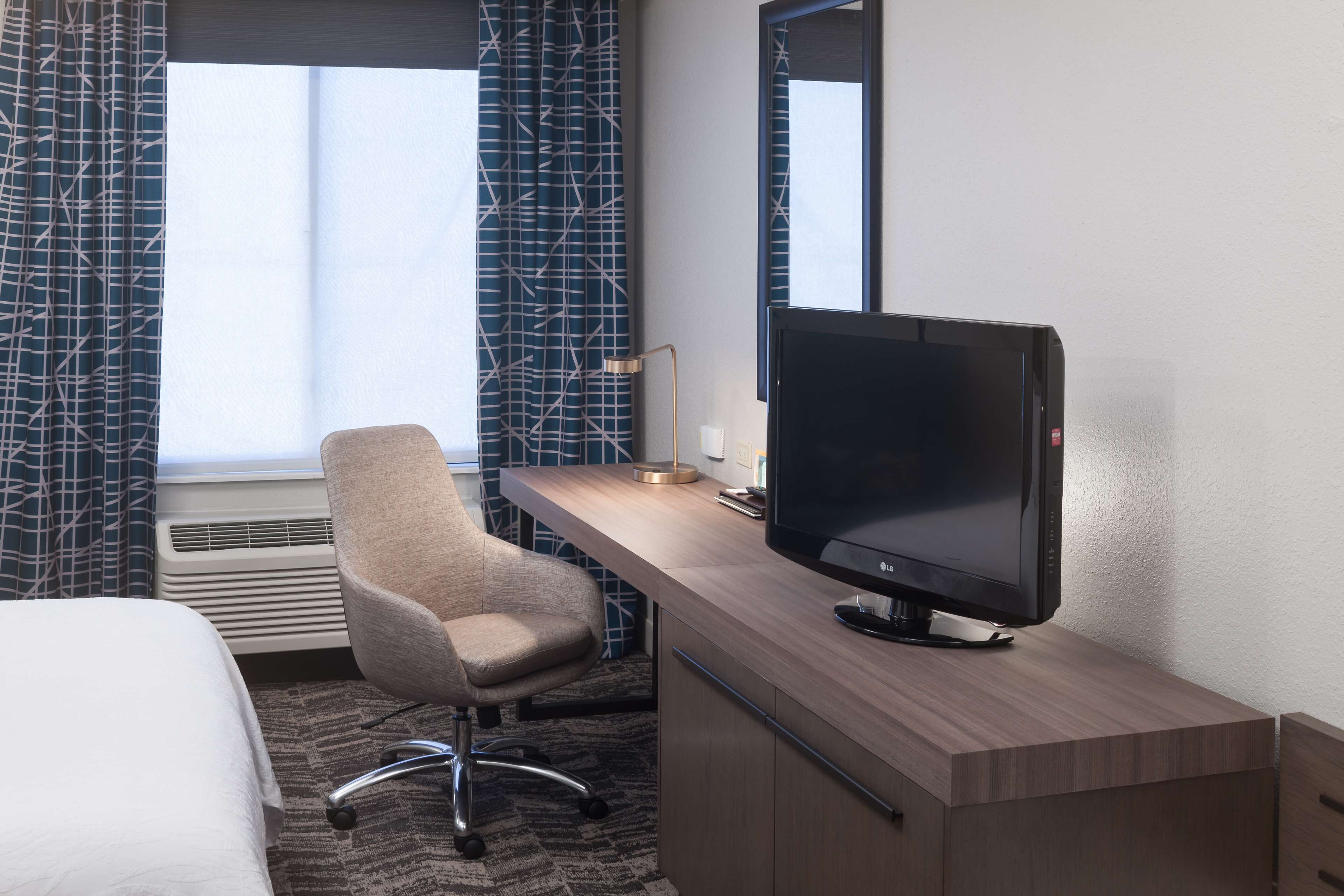 A hotel room with a bed, a desk with a television, a chair, and a window with patterned curtains.