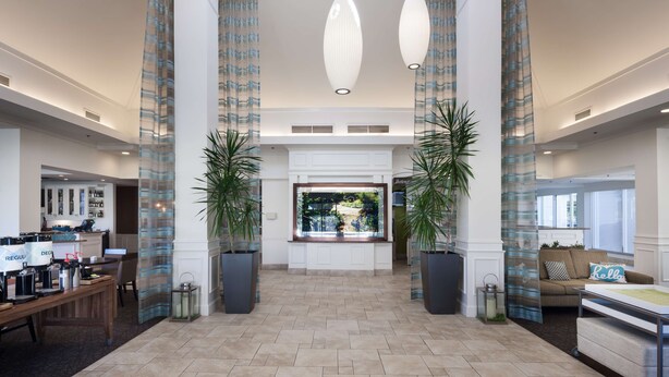 Een ruime lobby met een receptiebalie, twee grote potplanten, een zithoek met een bank en uitzicht door een groot raam.
