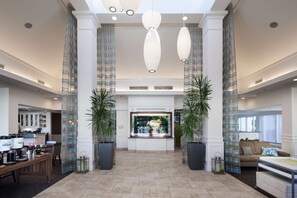 Un hall d’entrée spacieux avec un comptoir de réception, deux grands plants en pot, un coin salon avec un canapé et une vue dégagée sur l’extérieur grâce à une grande fenêtre.