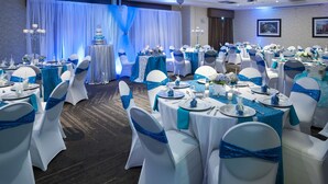 Een feestzaal met ronde tafels, witte stoelen met blauwe linten en een podium met een taart.