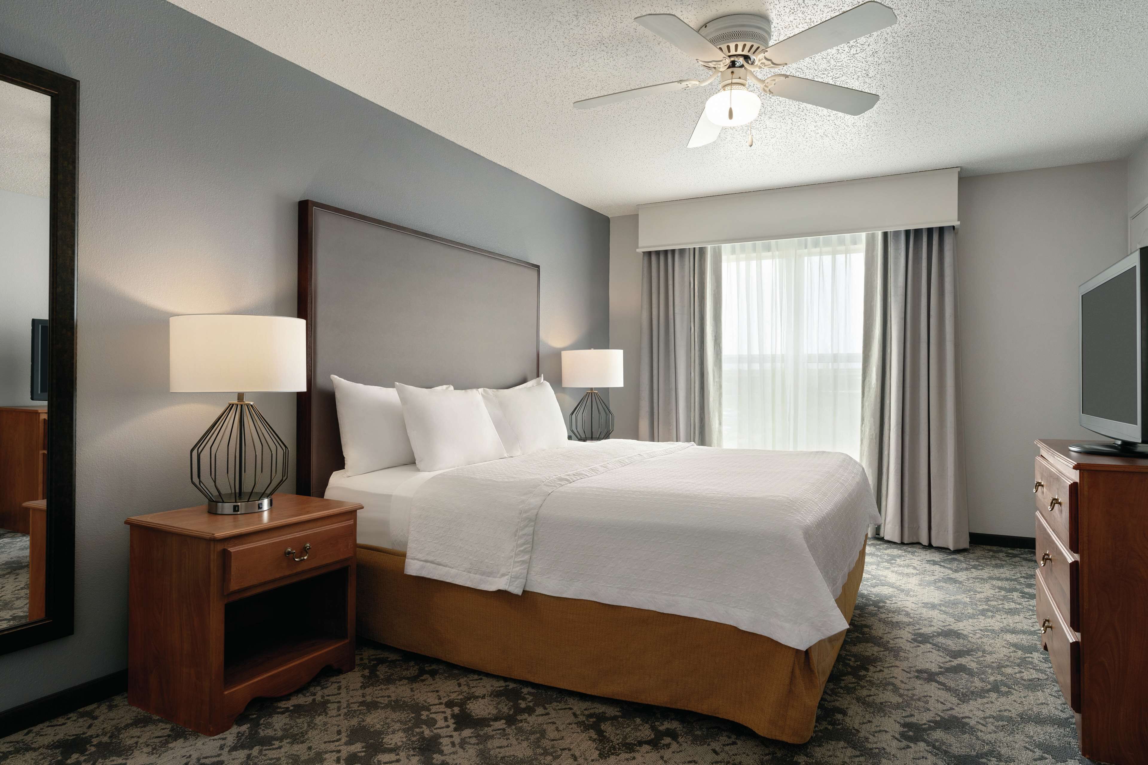 A hotel room with a large bed, two bedside tables with lamps, a ceiling fan, a TV, and a window with curtains.